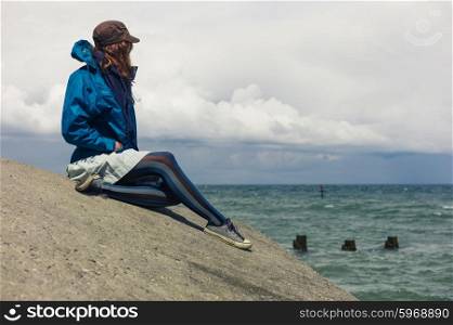 A young woman wearing a blue jacket is sitting on the flood defence by the sea