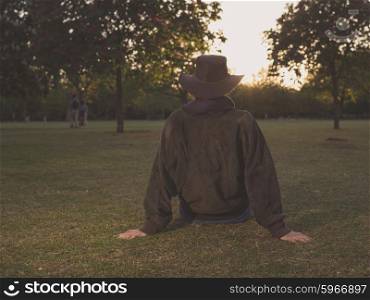 A young woman wearing a cowboy hat is sitting on the grass in a park watching the sunset