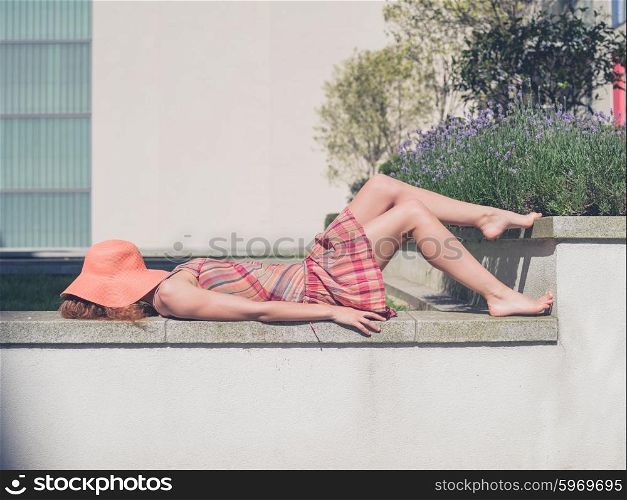 A young woman wearing a dress and a hat is relaxing in a green space outside an apartment building on a sunny summer day