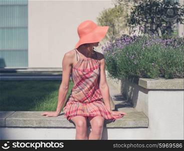 A young woman wearing a dress and a hat is relaxing outside an apartment building on the lawn by some lavender