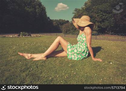 A young woman wearing a dress and a hat is sitting on the grass in a park