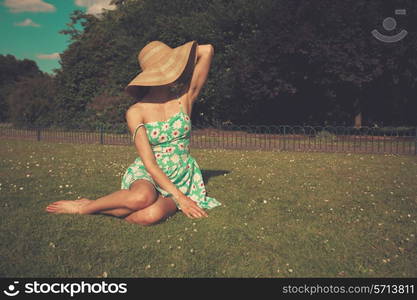 A young woman wearing a dress and a hat is sitting on the grass in a park