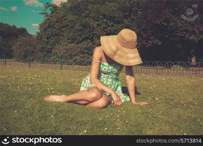 A young woman wearing a dress and a hat is sitting on the grass in a park