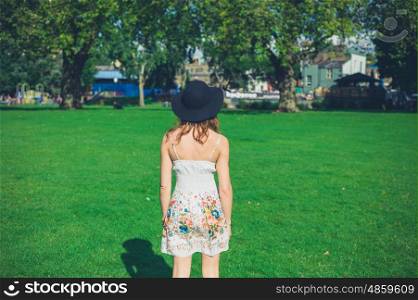 A young woman wearing a dress and a hat is standing on the grass in a park on a sunny summer day