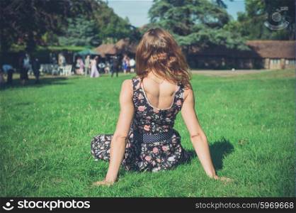 A young woman wearing a dress is relaxing on the grass on a sunny summer day and is watching people at a party in the distance