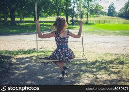 A young woman wearing a dress is sitting on a swing outside on a summer day
