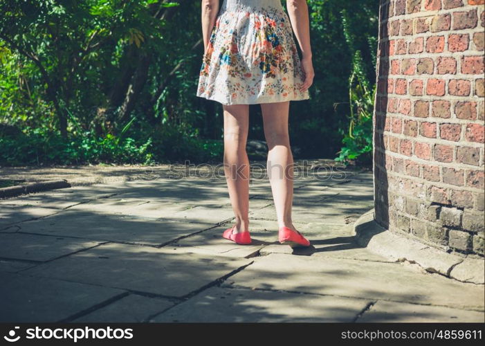 A young woman wearing a dress is standing by a brick structure in a park on a sunny summer day