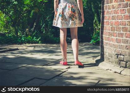 A young woman wearing a dress is standing by a brick structure in a park on a sunny summer day