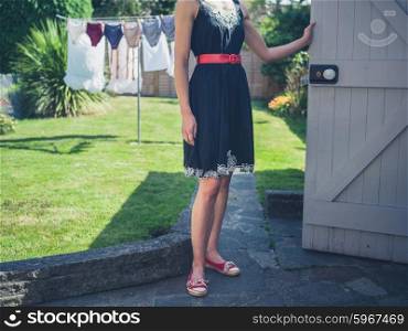 A young woman wearing a dress is standing by the door of a shed in a garden on a sunny summer day