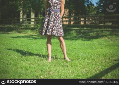 A young woman wearing a dress is standing in a field near a fence on a summer day
