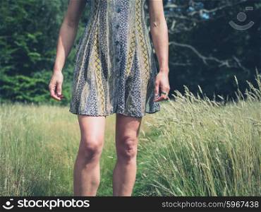 A young woman wearing a dress is standing in a meadow by a forest