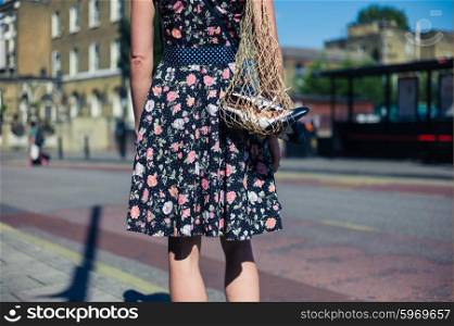 A young woman wearing a dress is standing in the street with a net bag containg a pair of spare party shoes