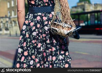 A young woman wearing a dress is standing in the street with a net bag containg a pair of spare party shoes