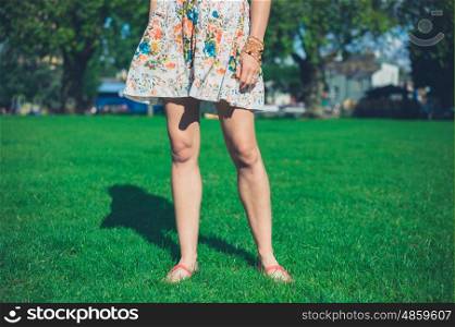 A young woman wearing a dress is standing on the grass in a park on a sunny summer day