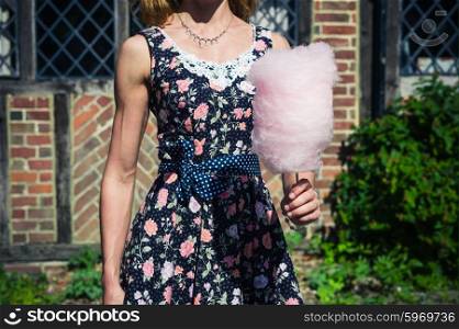 A young woman wearing a dress is standing outside an old farmhouse with a candyfloss on a sunny summer day