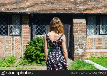A young woman wearing a dress is standing outside an old rustic country house on a sunny summer day