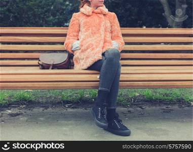 A young woman wearing a fur coat is sitting on a bench