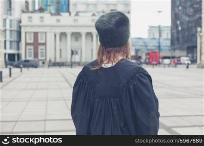 A young woman wearing a fur hat and a black graduation gown is standing in a square