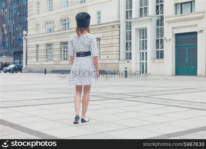 A young woman wearing a fur hat and a white dress with black spots is walking in a square