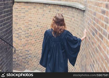 A young woman wearing a graduation gown is walking down some stairs outside by a brick wall