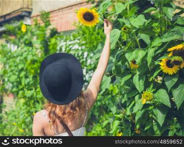 A young woman wearing a hat and a dress is admiring some sunflowers outside a house on a sunny day in summer