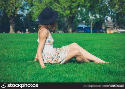 A young woman wearing a hat and a dress is sitting on the grass in a park on a sunny summer day