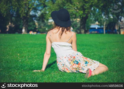 A young woman wearing a hat and a dress is sitting on the grass in a park on a sunny summer day