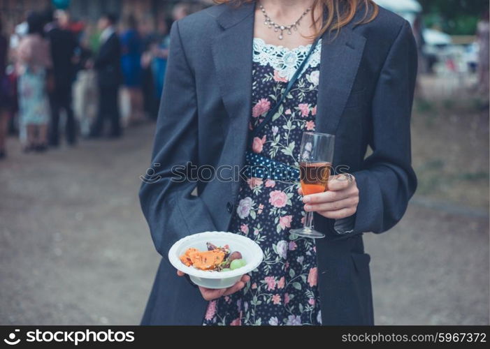 A young woman wearing a man&rsquo;s jacket over her dress is standing with a drink at a party in the countryside