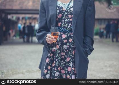 A young woman wearing a man&rsquo;s jacket over her dress is standing with a drink at a party in the countryside