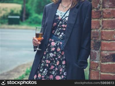 A young woman wearing a man&rsquo;s jacket over her dress is standing with a drink by a wall in the countryside