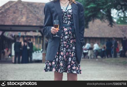 A young woman wearing a man&rsquo;s jacket over her dress is standing with a drink at a party in the countryside