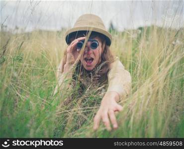A young woman wearing a safari hat is hiding in the tall grass and is looking through binoculars