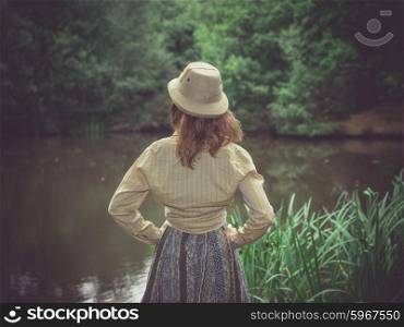 A young woman wearing a safari hat is standing by a pond in the forest on a sunny summer day