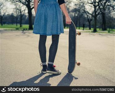 A young woman wearing a skirt is standing in a park with a skateboard