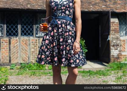A young woman wearing a stylish dress is standing outside a rural country house with a drink in her hand on a sunny day