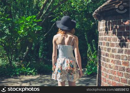 A young woman wearing a summer dress and a black hat is walking in a park by an old brick structure