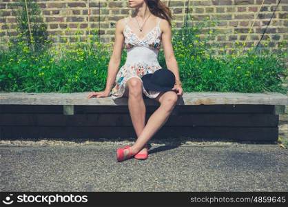 A young woman wearing a summer dress is sitting on a bench outside with a hat