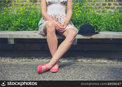 A young woman wearing a summer dress is sitting on a bench outside with a hat