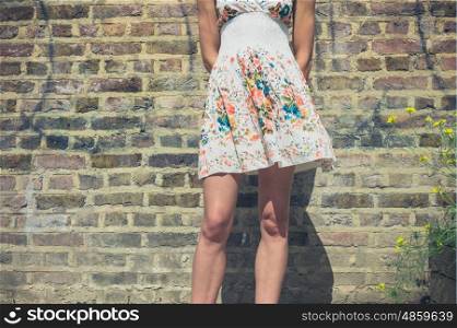 A young woman wearing a summer dress is standing by a brick wall outside on a sunny day