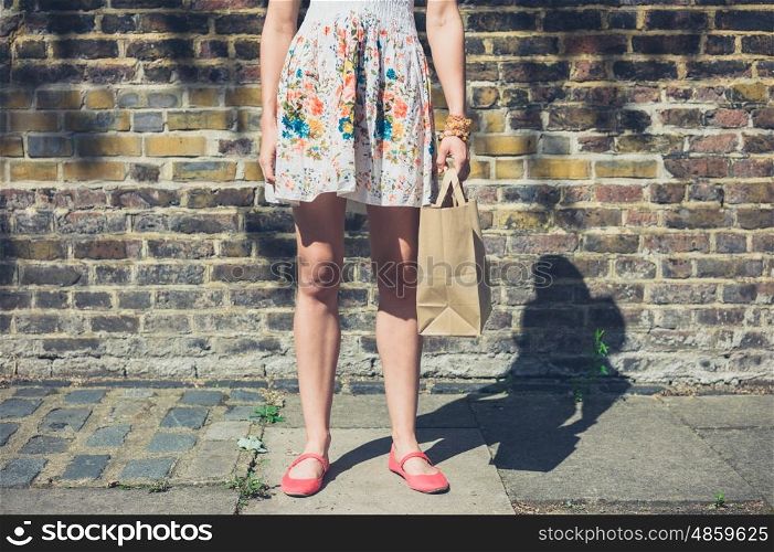 A young woman wearing a summer dress is standing in the street with a paper bag