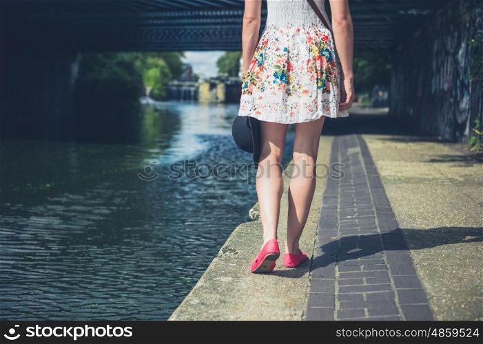 A young woman wearing a summer dress is walking by the canal with a hat in her hand