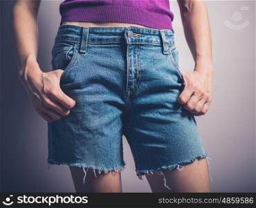 A young woman wearing denim shorts is posing with her hands in her pockets