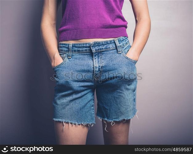 A young woman wearing denim shorts is posing with her hands in her pockets