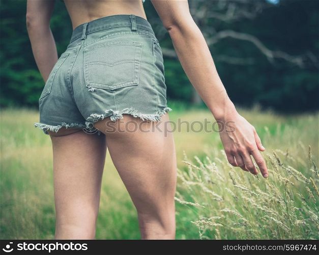 A young woman wearing short shorts is touching the grass as she is walking in a meadow in the forest
