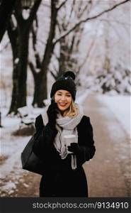 A young woman wearing warm winter clothes and a knit hat smiles happily as she stands in the snow and using mobile phone whil holding cofee cup