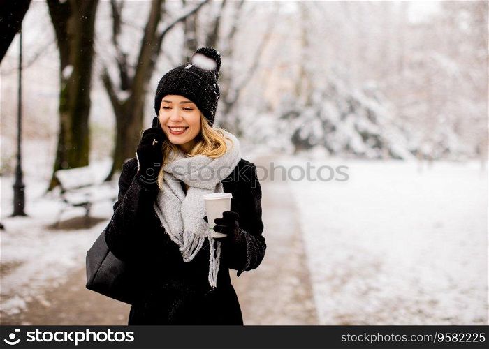 A young woman wearing warm winter clothes and a knit hat smiles happily as she stands in the snow and using mobile phone whil holding cofee cup