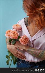 A young woman with a tattoo holds in her hand a bouquet of pink roses around a blue background with copy space. Mother's day concept. A girl with a tattoo on her hand holds delicate pink roses around a blue background with space for text. Valentine's Day Gift