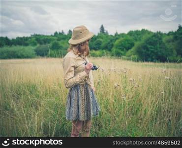 A young woman with binoculars and a safari hat is standing in a field in the wilderness