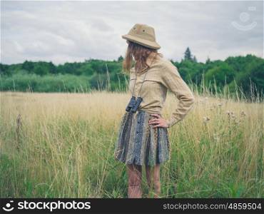 A young woman with binoculars and a safari hat is standing in a field in the wilderness