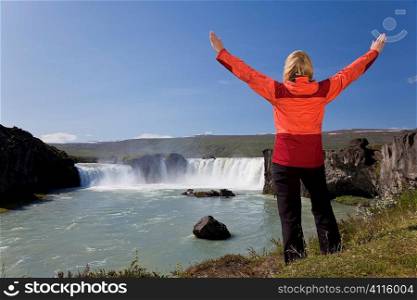A young woman with her arms up in celebration of achievement by a waterfall. Shot on location at Godafoss waterfall in Iceland.